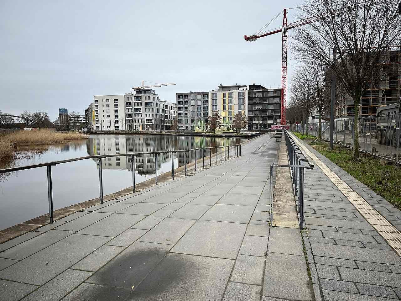 A paved promenade along a still harbour basin in the Neckarbogen district, with new apartment buildings across the water and a construction crane