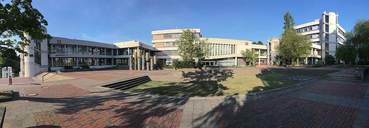 Panorama of the University of Hildesheim main campus: low concrete lecture buildings around a brick-paved square with the Audimax on the left