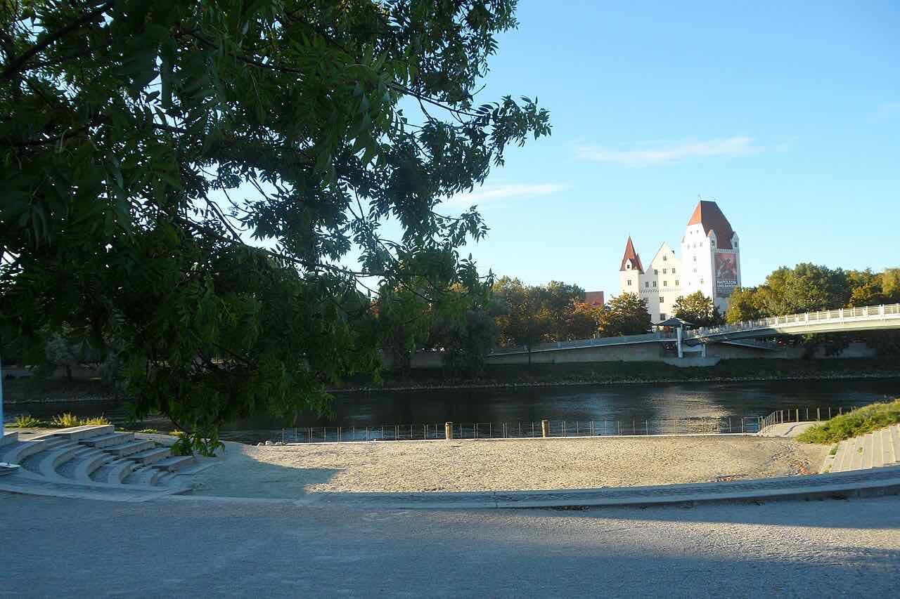 A gravel beach and stone steps on the bank of the Danube in Ingolstadt, with the Neues Schloss and a footbridge across the river in the background
