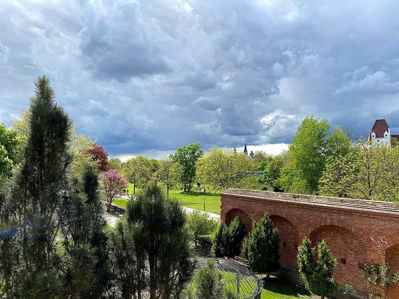 Green lawns and trees in the Klenzepark with a long red brick wall with arches on the right, a storm cloud overhead and the castle roof at the edge of the picture