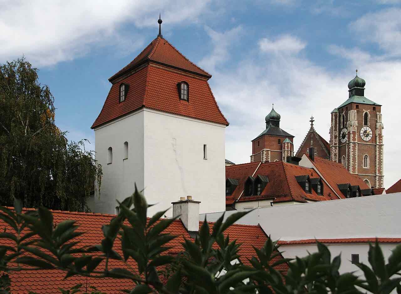 A white medieval tower with a steep red roof beside red-tiled roofs, with the brick towers of the Liebfrauenmünster rising behind