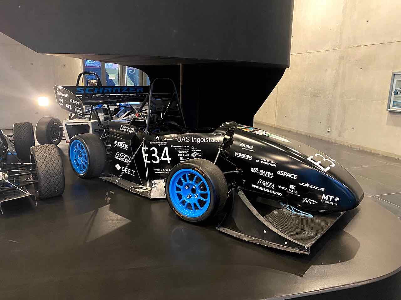 A black Formula Student race car with blue wheels and the words Schanzer and UAS Ingolstadt on its body, shown in a concrete exhibition hall