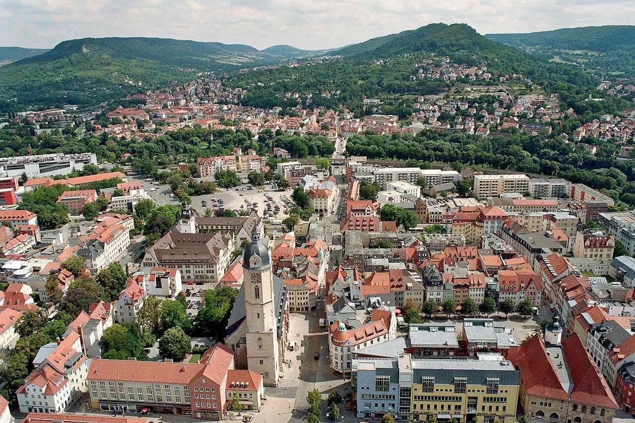 Aerial view over the old town of Jena with a church tower, red roofs and tree-covered hills rising on both sides of the valley