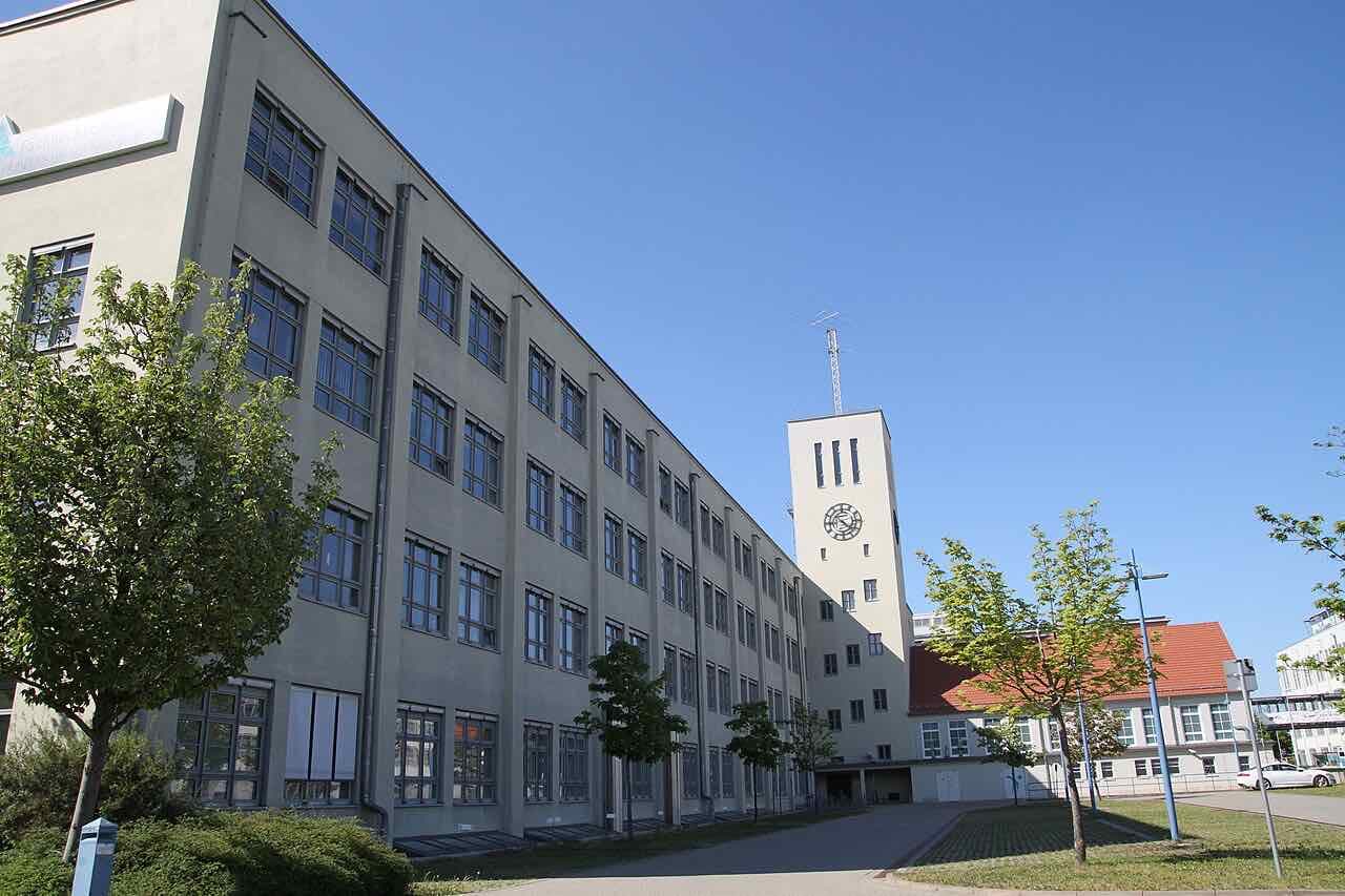 A long pale building of the Ernst-Abbe-Hochschule campus with tall windows and a clock tower, under a clear blue sky
