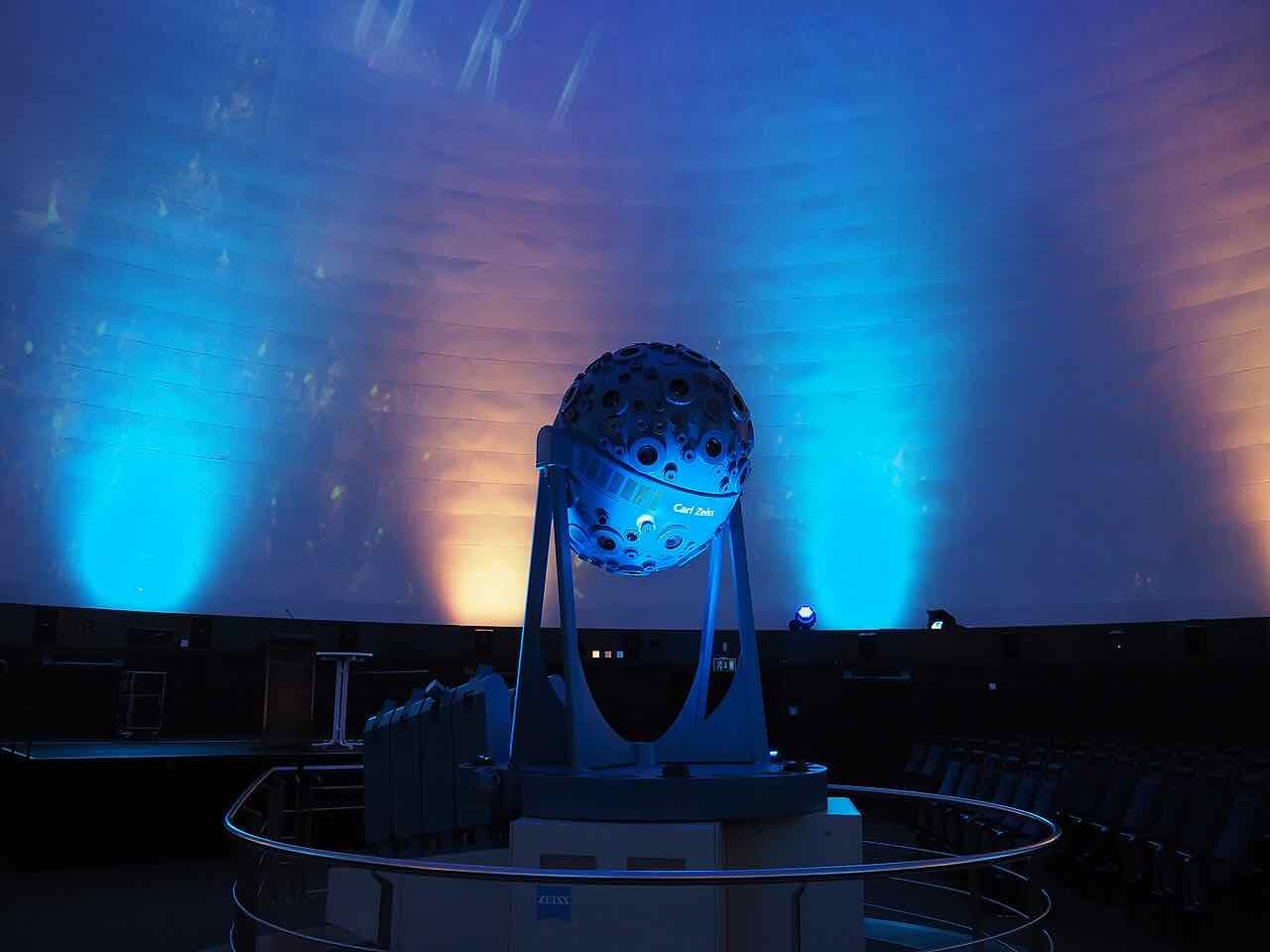 A Zeiss star projector, a dark ball on a stand, in front of a dome lit blue and orange inside the planetarium in Jena
