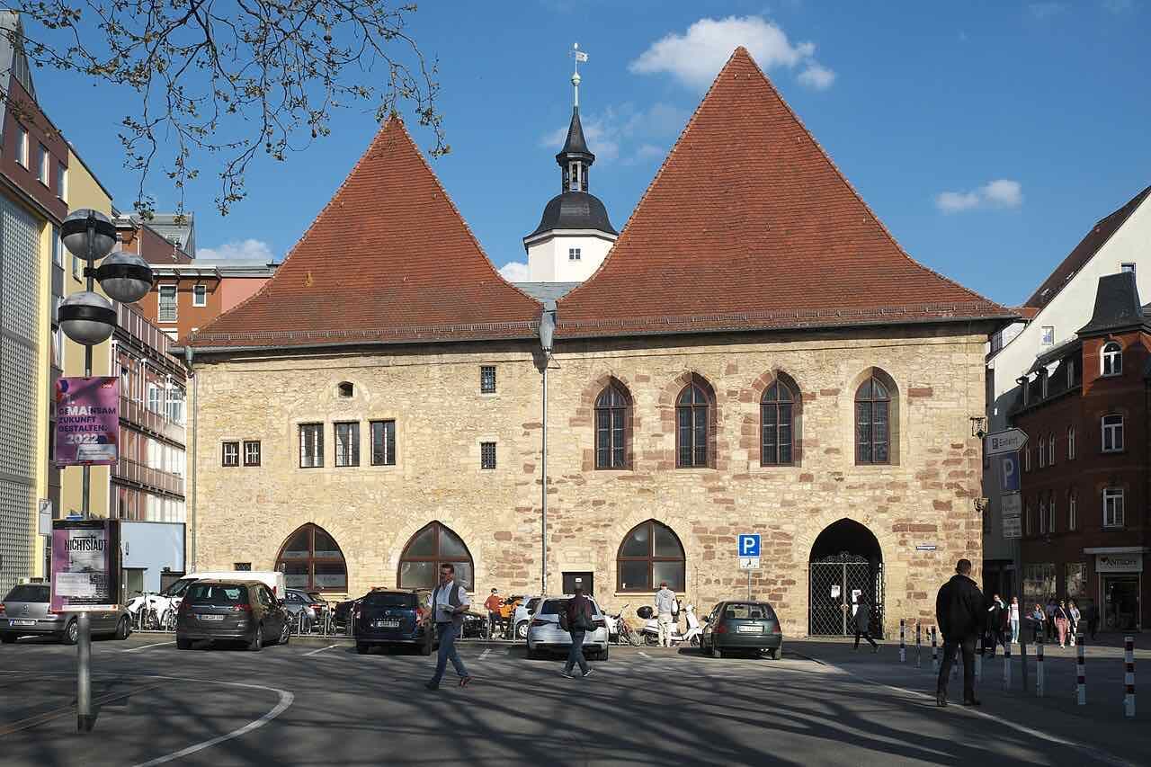 The old town hall of Jena, a stone building with two steep red gables and a small tower behind, with people crossing the square in front