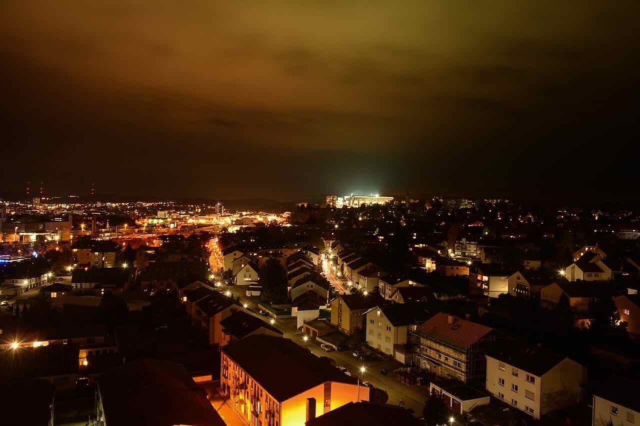 Kaiserslautern at night seen over rooftops, with the floodlit Fritz-Walter-Stadion glowing on the hill in the distance