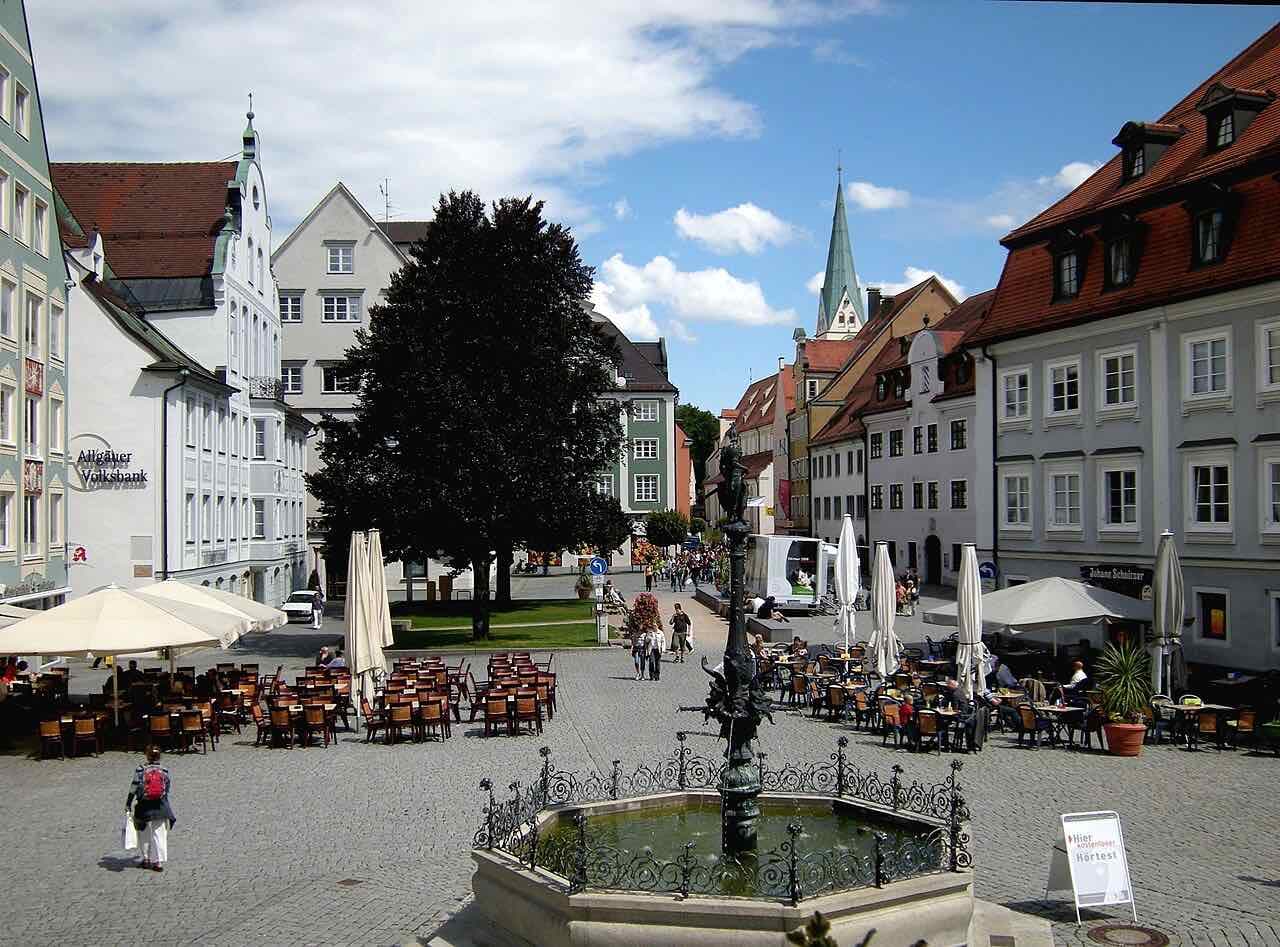 The Rathausplatz in Kempten on a sunny day, with a wrought-iron fountain in front, cafe tables under parasols and a green church spire above the gabled town houses
