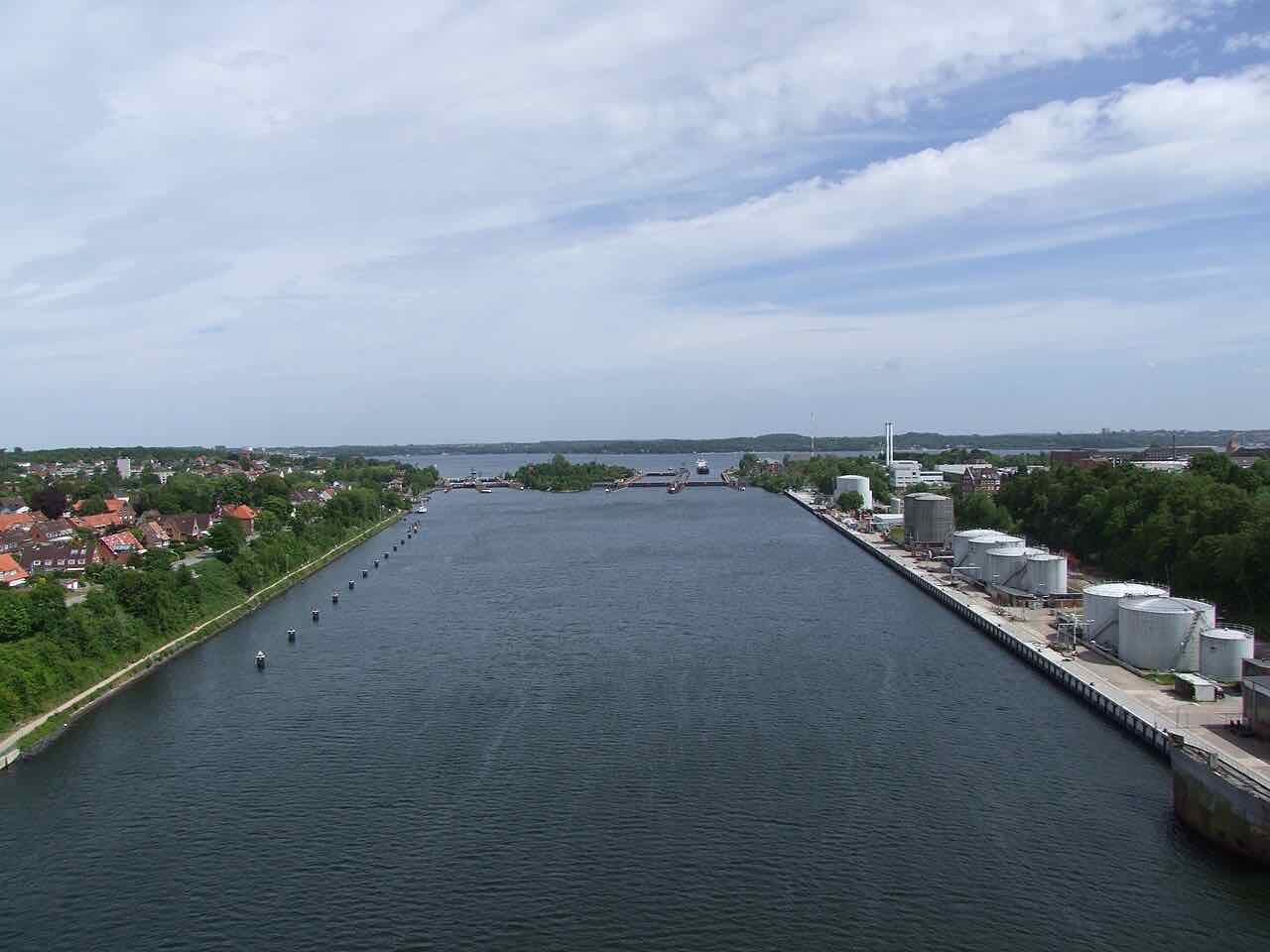The Kiel Canal running straight between green banks towards the Kiel Fjord, with fuel tanks on the right bank
