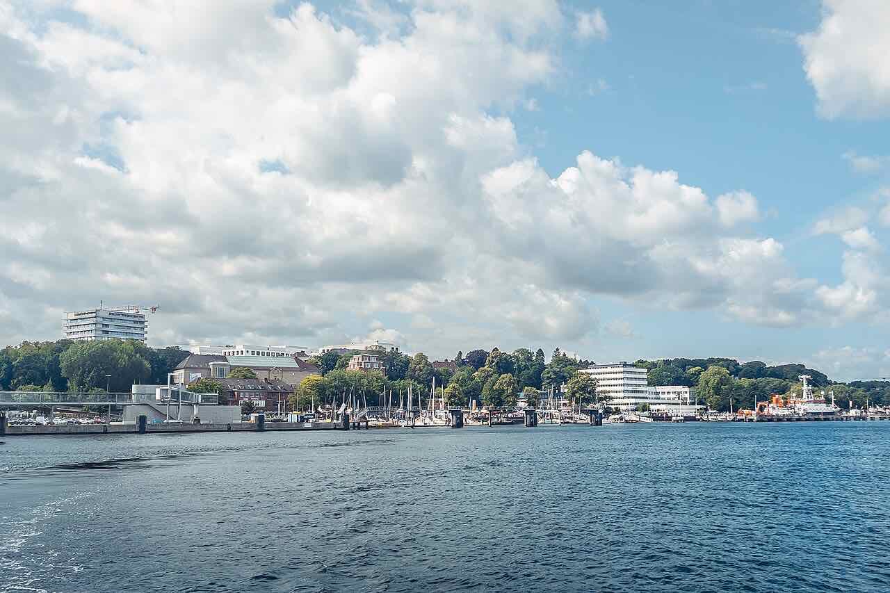 The Kiel waterfront and the Kiellinie seen from the water under a big cloudy sky, with sailing boats in the marina