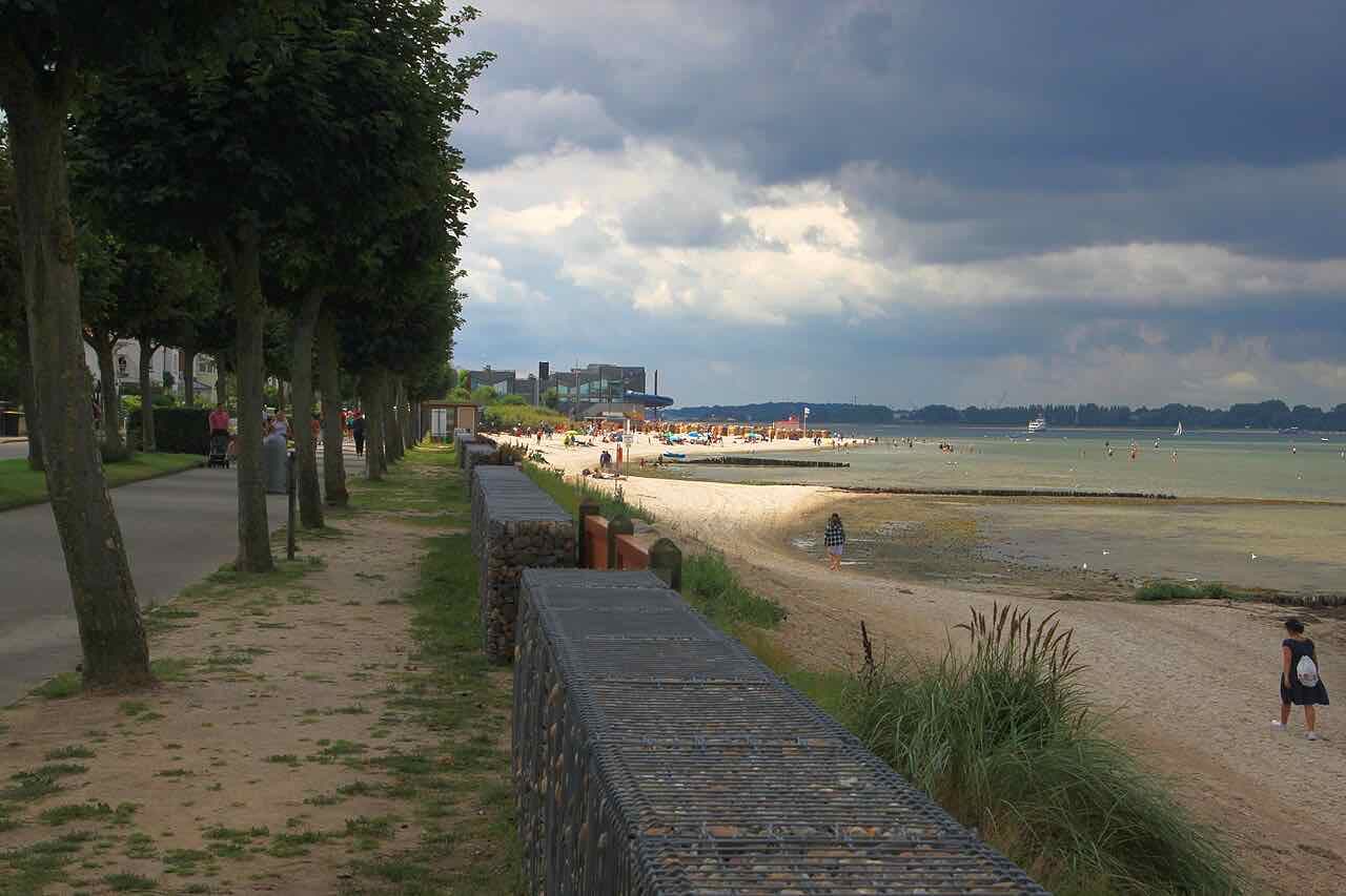 A sandy beach and a tree-lined promenade at Laboe under a stormy sky, with a few walkers