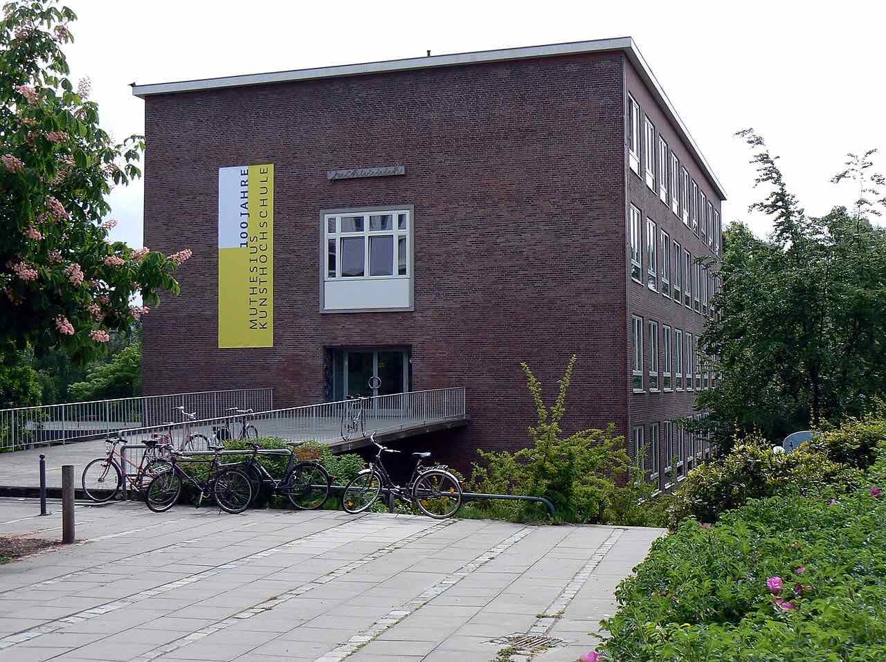 A red-brick building of the Muthesius Kunsthochschule with a yellow banner, bicycles and flowering shrubs in front