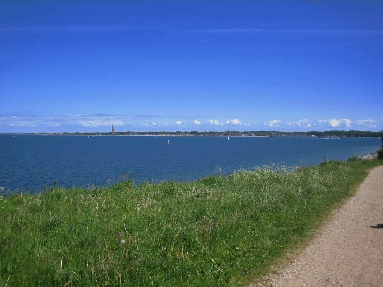 A grassy dyke and a path above the blue Kiel Fjord at Schilksee with the Laboe shore on the horizon