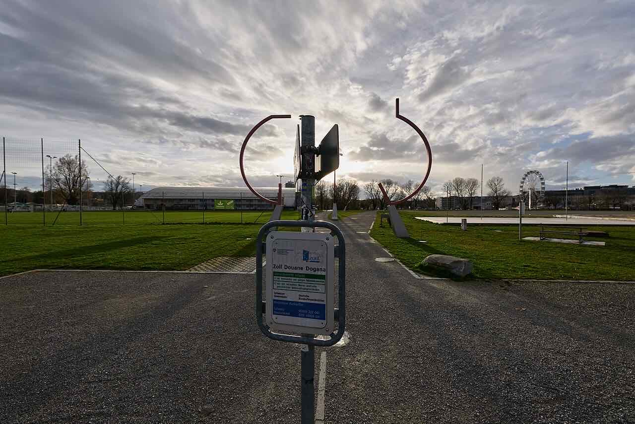 A Swiss customs sign on a post in an open green space in Konstanz, with a red sculpture and a small ferris wheel in the distance