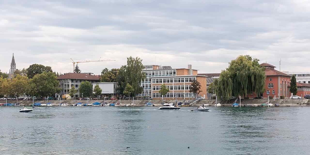 HTWG Konstanz buildings on the bank of the Rhine, with moored boats in front and the Münster spire among trees on the left