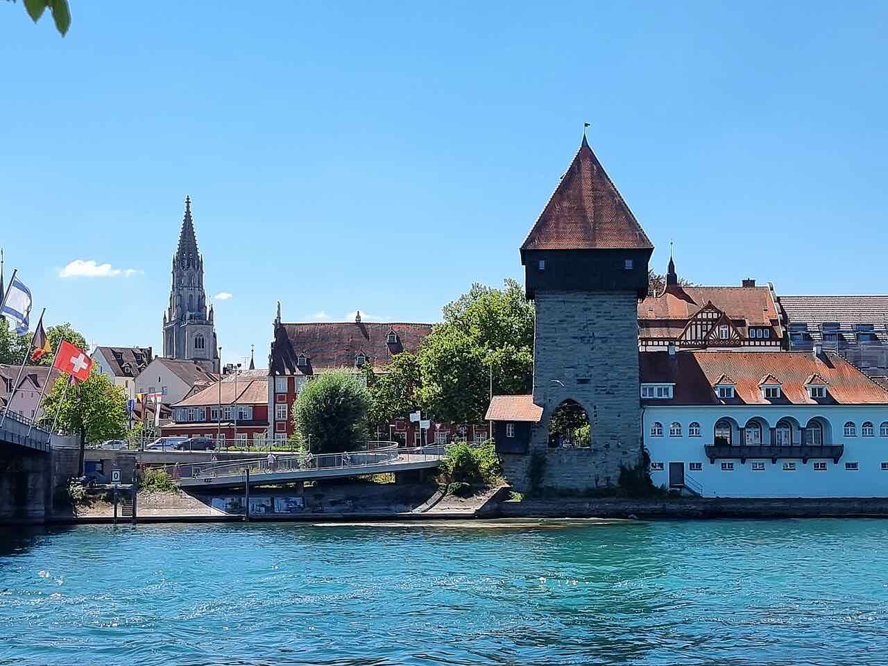 The Rheintorturm, a stone tower with a steep red roof, beside the Rhine with the Münster spire and a Swiss flag in the background