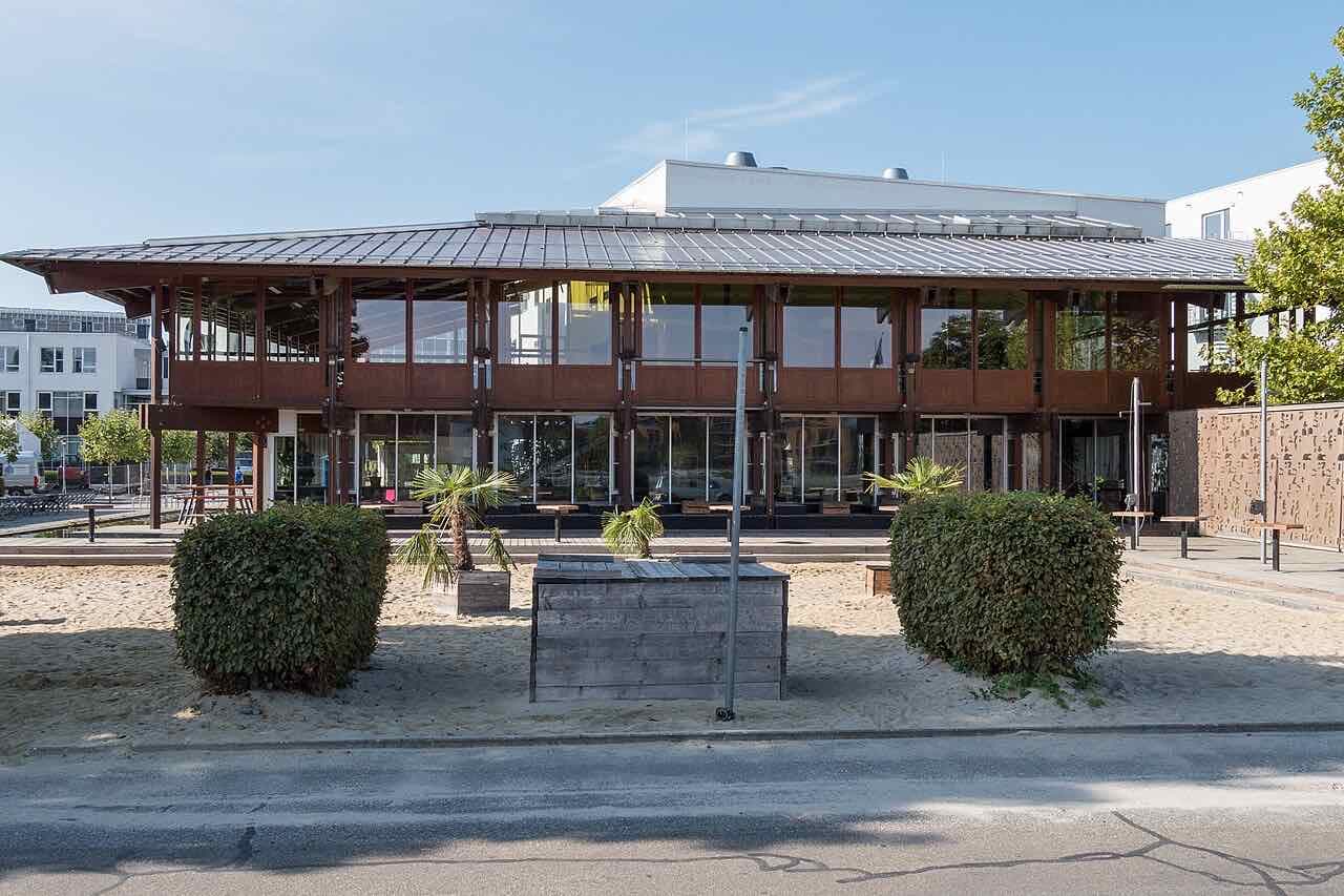 The wood-and-glass beach bar building of HTWG Konstanz standing on a sand area with palm trees and benches