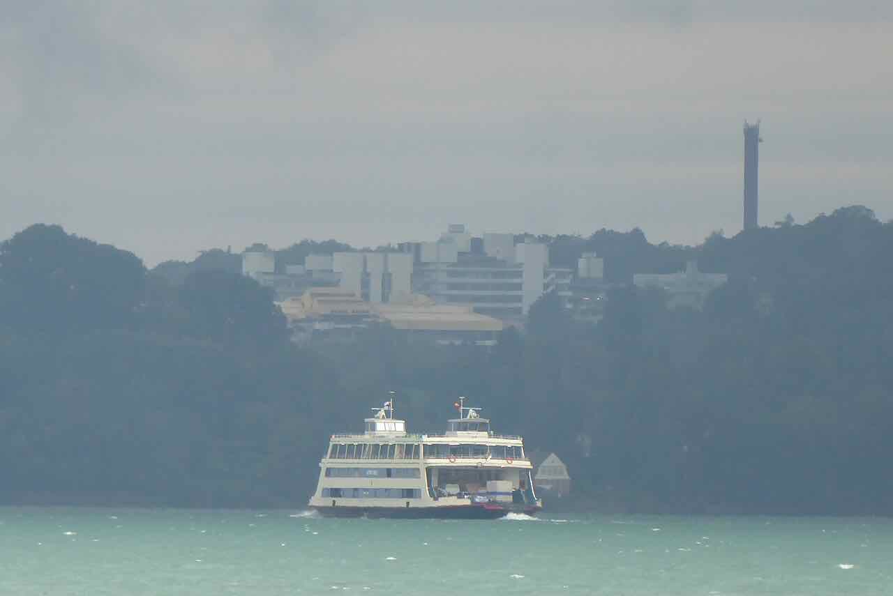 A Konstanz car ferry on the lake with the pale university buildings and a tall tower on the wooded hillside behind it