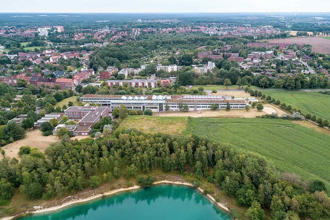 Aerial view of the Leuphana Volgershall campus buildings between fields, trees and the turquoise Kalkbruchsee quarry lake, with the town behind
