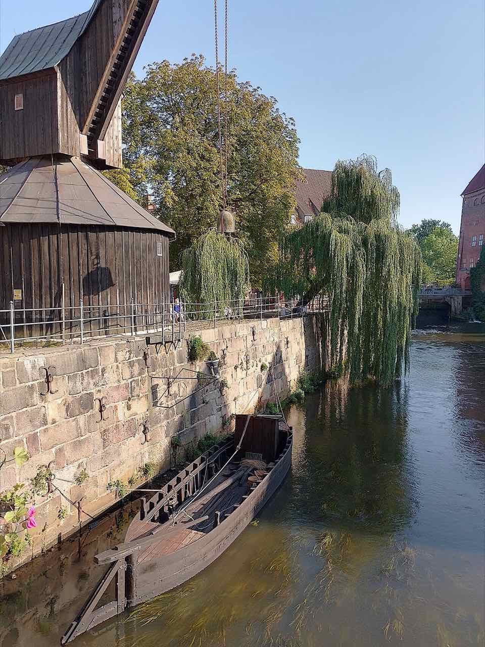 The wooden harbour crane beside the Ilmenau with a historic wooden river boat moored under the stone quay wall and weeping willows