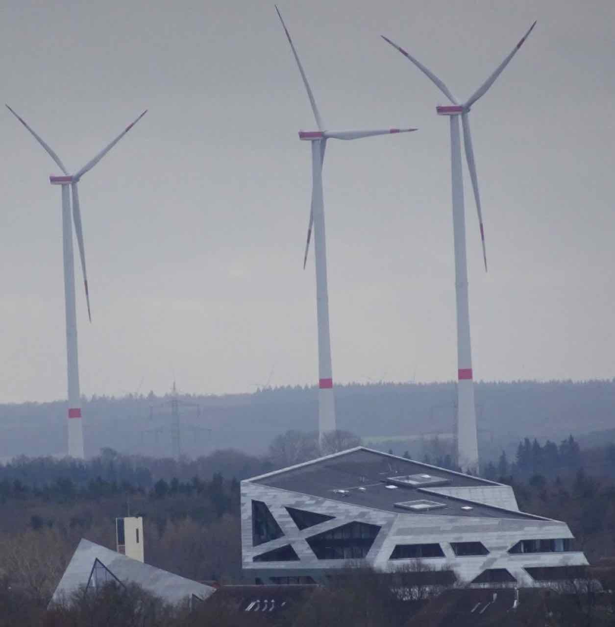 The angular silver-grey Leuphana Central Building seen from the water tower, with three wind turbines and forest behind it under a grey sky