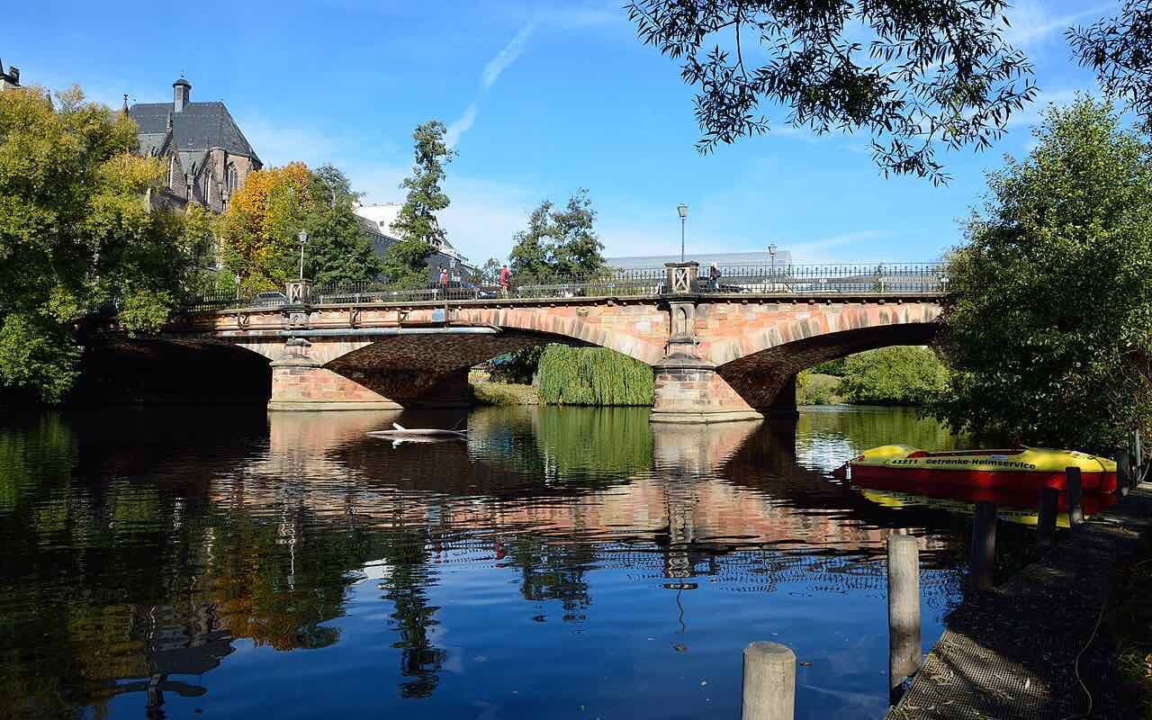 The Weidenhäuser Brücke, a red sandstone arched bridge over the calm Lahn, with a rowing boat at the right and a church roof behind the trees