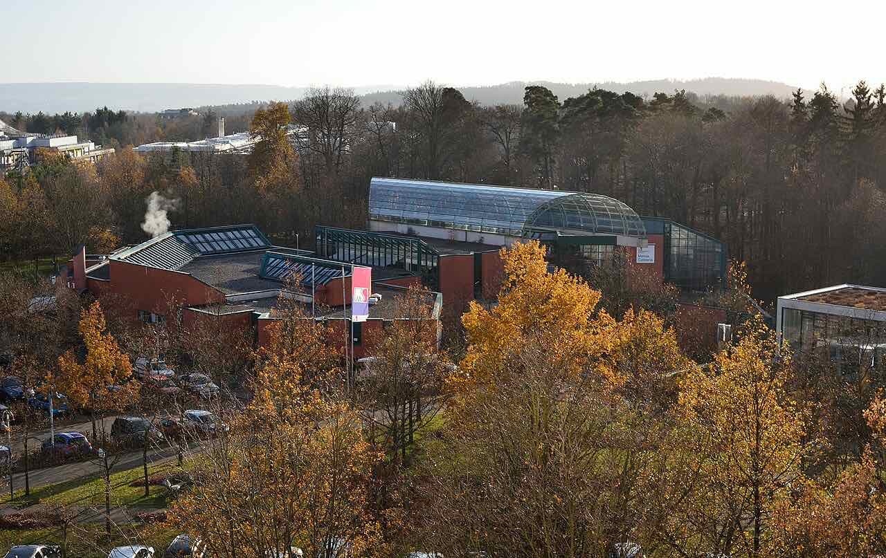 The Lahnberge campus in autumn: the glass-roofed Mensa and red brick buildings among yellow trees, with forest behind
