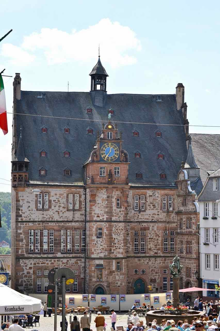 The town hall on the market square, a stone building with a steep slate roof, a clock and a small tower, with people on the square below