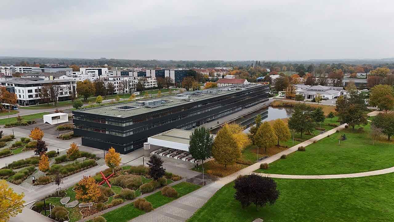 Aerial view of the Hochschule Neu-Ulm campus, a long dark building beside a pond and landscaped gardens in autumn colours