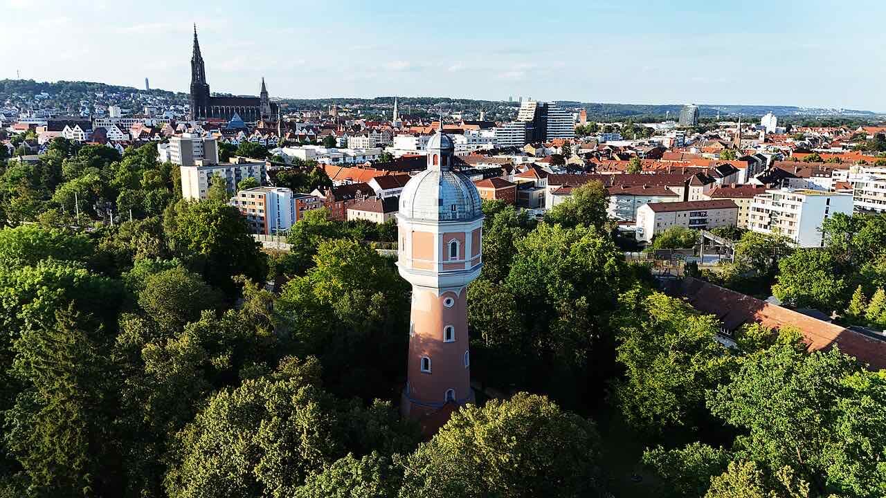 The pink Neu-Ulm water tower rising out of green trees, with the red roofs of the city and the tall spire of Ulm Minster in the background
