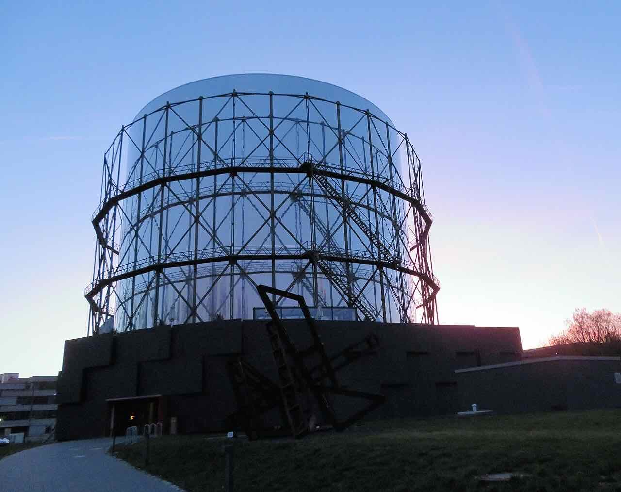 The Gasometer at dusk, its glass-and-steel drum lit by a pink and blue sky, with a dark steel sculpture on the lawn in front