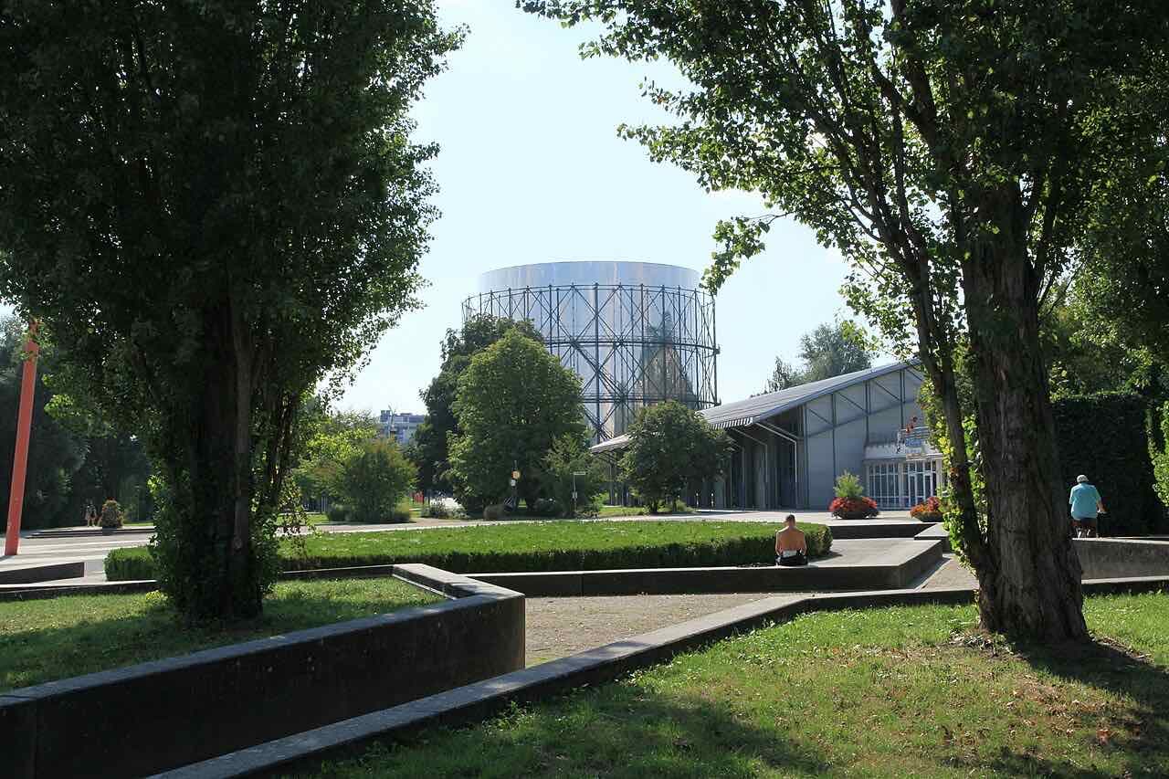 The Pforzheim Gasometer, a round steel-framed tower with a silver top, seen across lawns and trees in the Enzauenpark on a sunny day