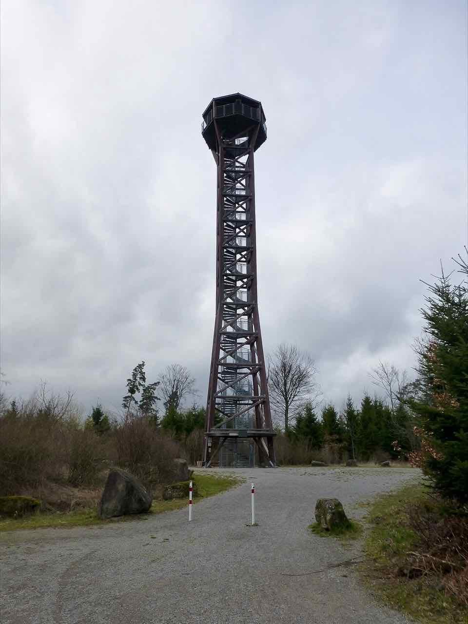 The tall wooden Hohe Warte lookout tower on a gravel clearing in the forest under a grey sky