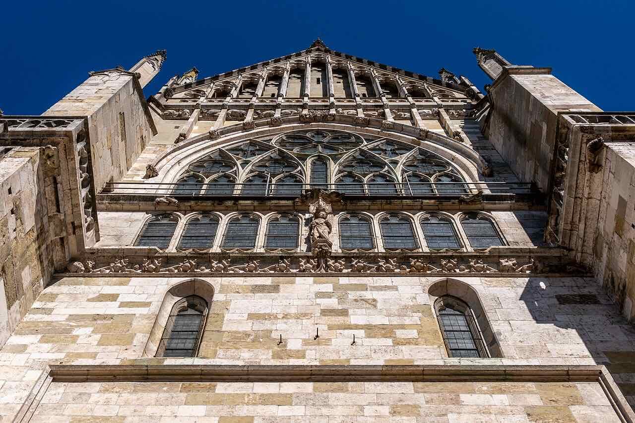 The Gothic stonework of Regensburg Cathedral seen from below, with traceried windows and a large round window against a deep blue sky