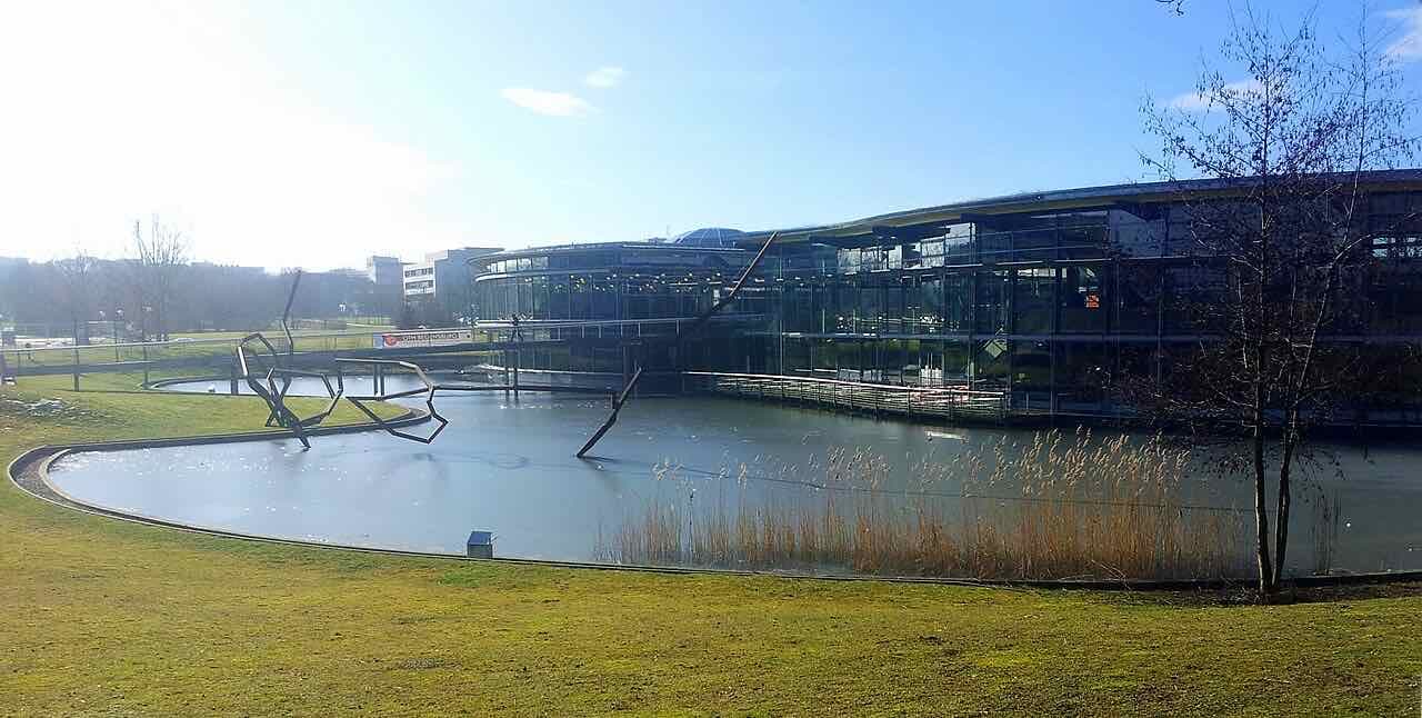 A glass-fronted building beside a pond with a metal sculpture and reeds on the OTH Regensburg campus