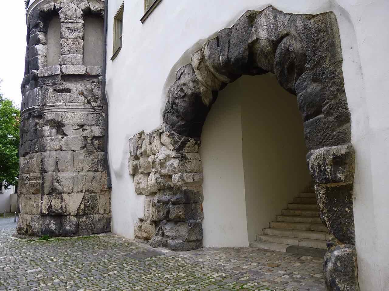 The Roman Porta Praetoria: a dark stone arch and a round tower built into the wall of a white house, with a stair behind the arch