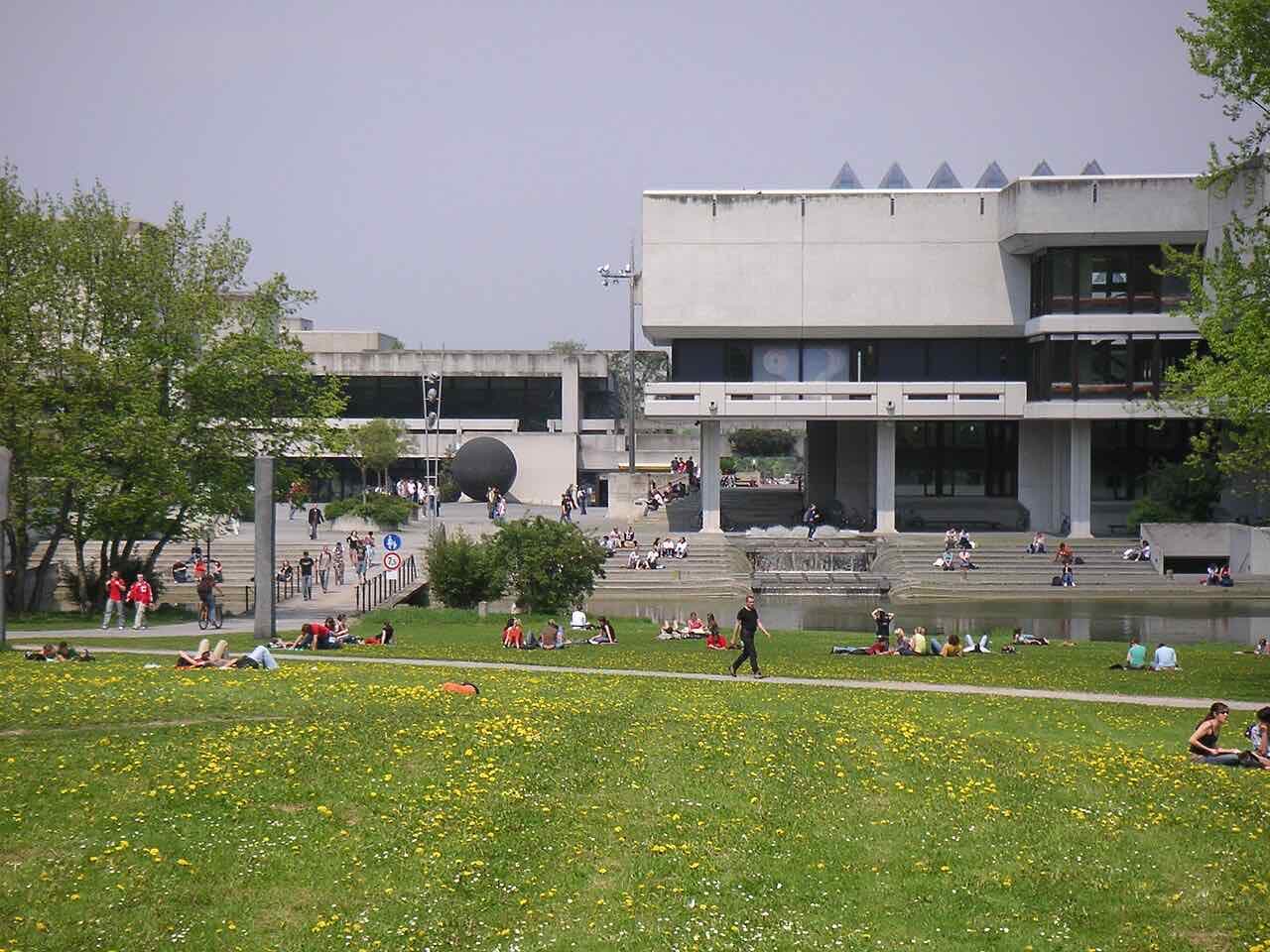 Students lying on a spring lawn in front of the University of Regensburg library and a pond, with a large dark sphere sculpture behind