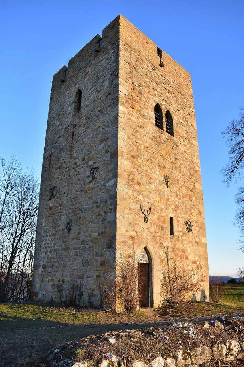 A square stone tower with small arched windows and a wooden door on a grassy hilltop in low evening light