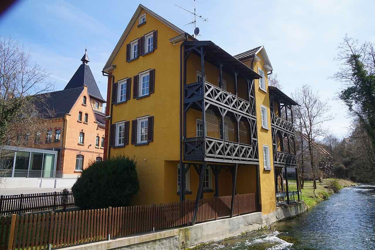A yellow house with dark wooden balconies beside the fast-flowing Echaz, with a red-brick building with a pointed tower behind it