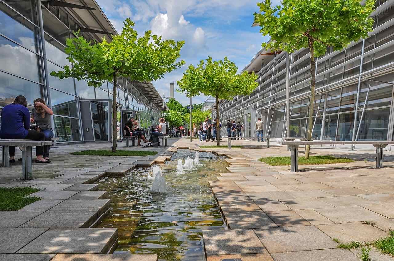 The paved campus square between two glass buildings at TH Rosenheim, with a water channel, benches, young trees and students sitting and walking