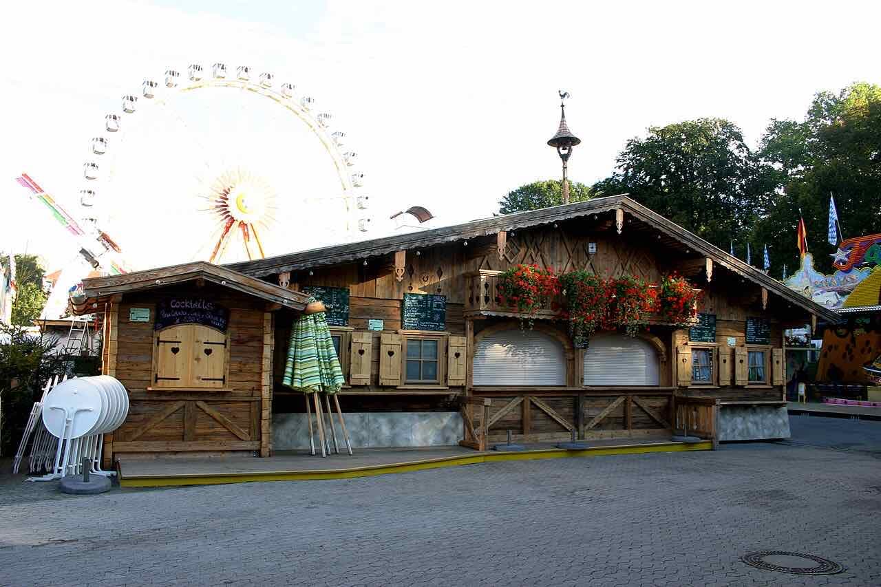 A wooden festival chalet with flower boxes and a large Ferris wheel behind it on a fairground in Rosenheim