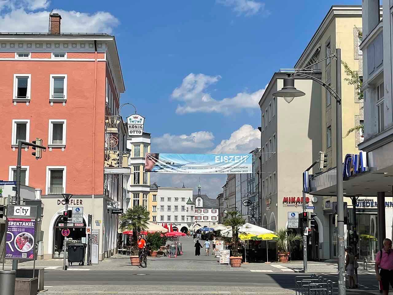 Looking along a pedestrian street in Rosenheim towards the old town square, with cafe umbrellas, colourful town houses and the town hall tower under a blue sky
