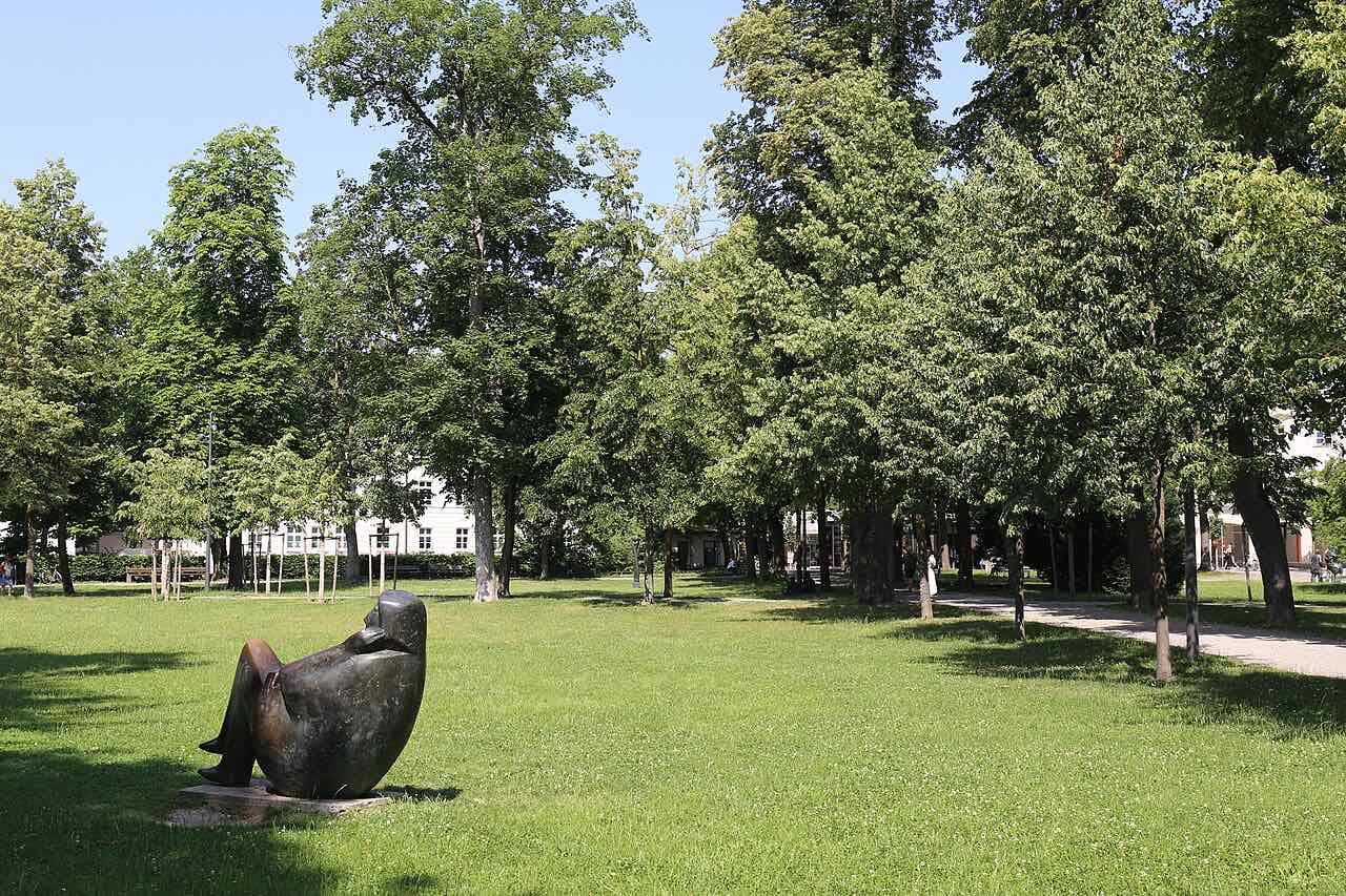 A bronze sculpture of a seated figure on a lawn among tall lime trees in the Salingarten park in Rosenheim