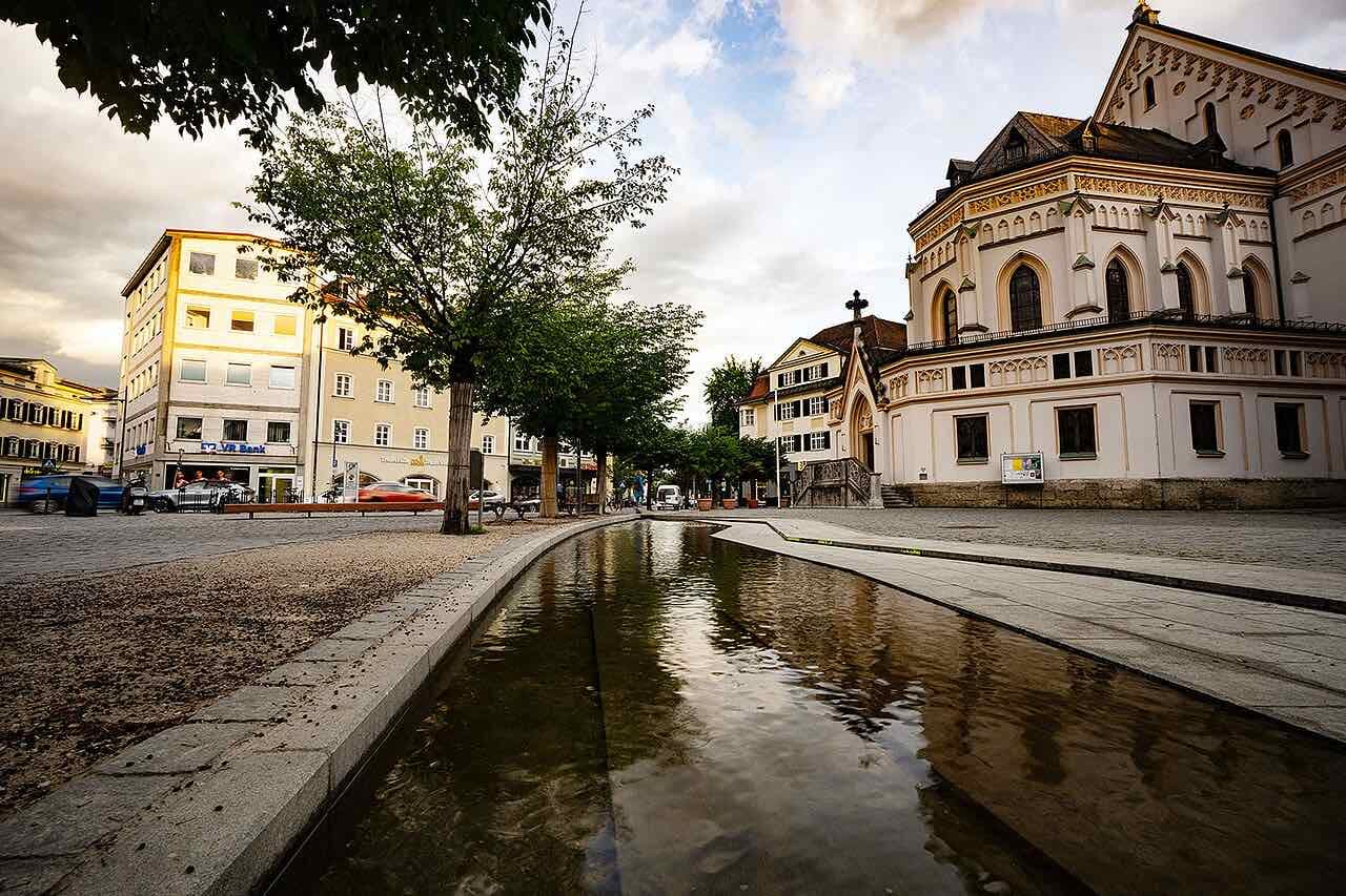 A narrow stone-lined water channel running across a square towards the cream-coloured neo-Gothic Erlöserkirche in Rosenheim