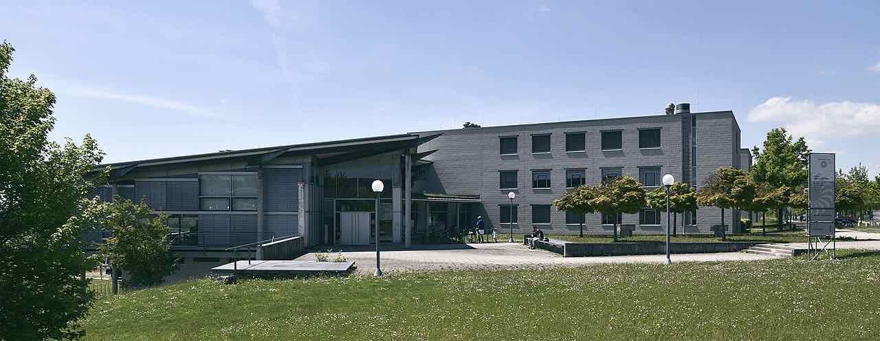 A low grey university building with a pitched entrance roof and a lawn in front, on the Albert-Einstein-Allee campus