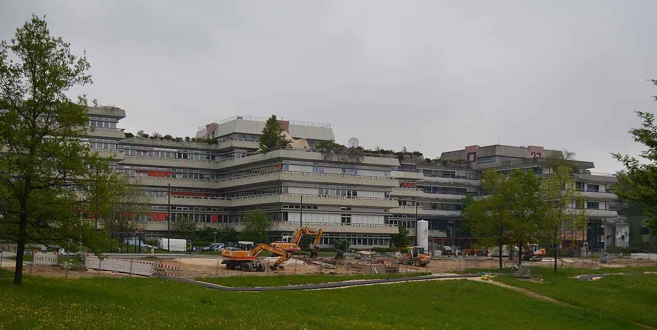 A long terraced concrete university building on a green slope under a grey sky, with building work in the foreground