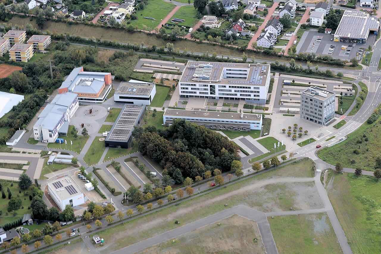 Aerial view of the OTH Amberg-Weiden campus in Weiden: several white and grey university buildings with car parks, lawns and trees, and houses beyond
