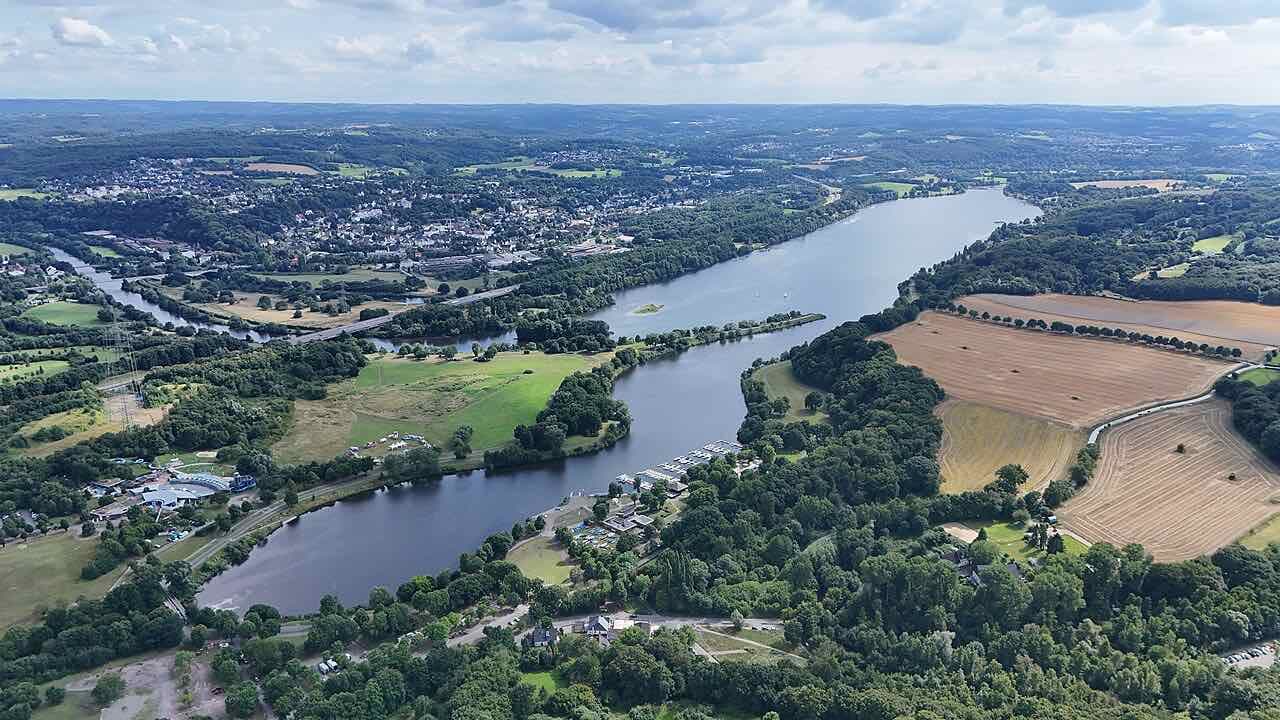 An aerial view of a long reservoir winding between green woods and fields, with the rooftops of a town on the far shore and hills on the horizon