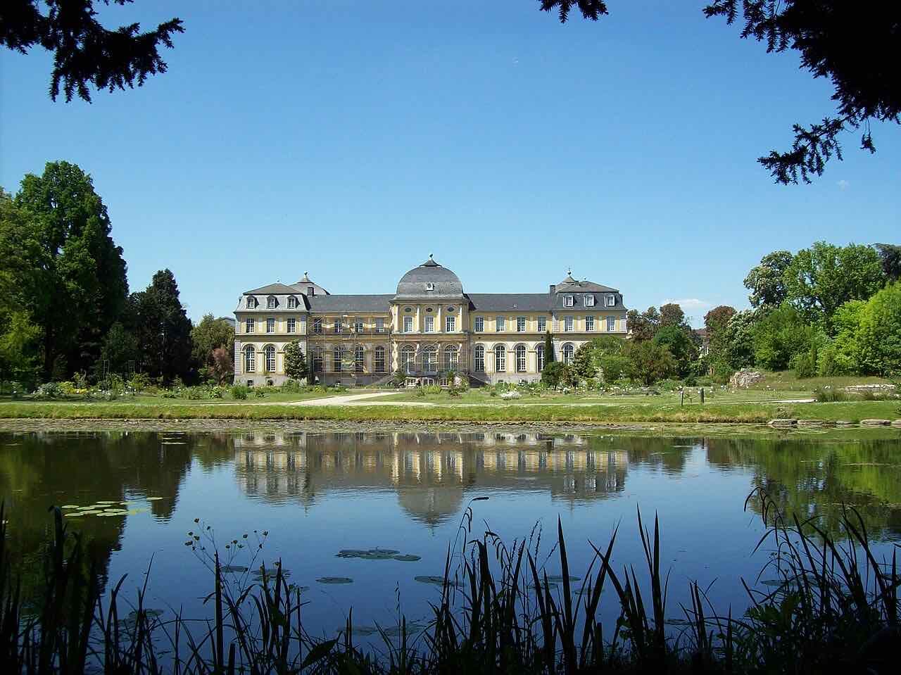 Poppelsdorf Palace, a pale yellow palace with a domed centre, reflected in a pond in the botanical garden under a clear blue sky