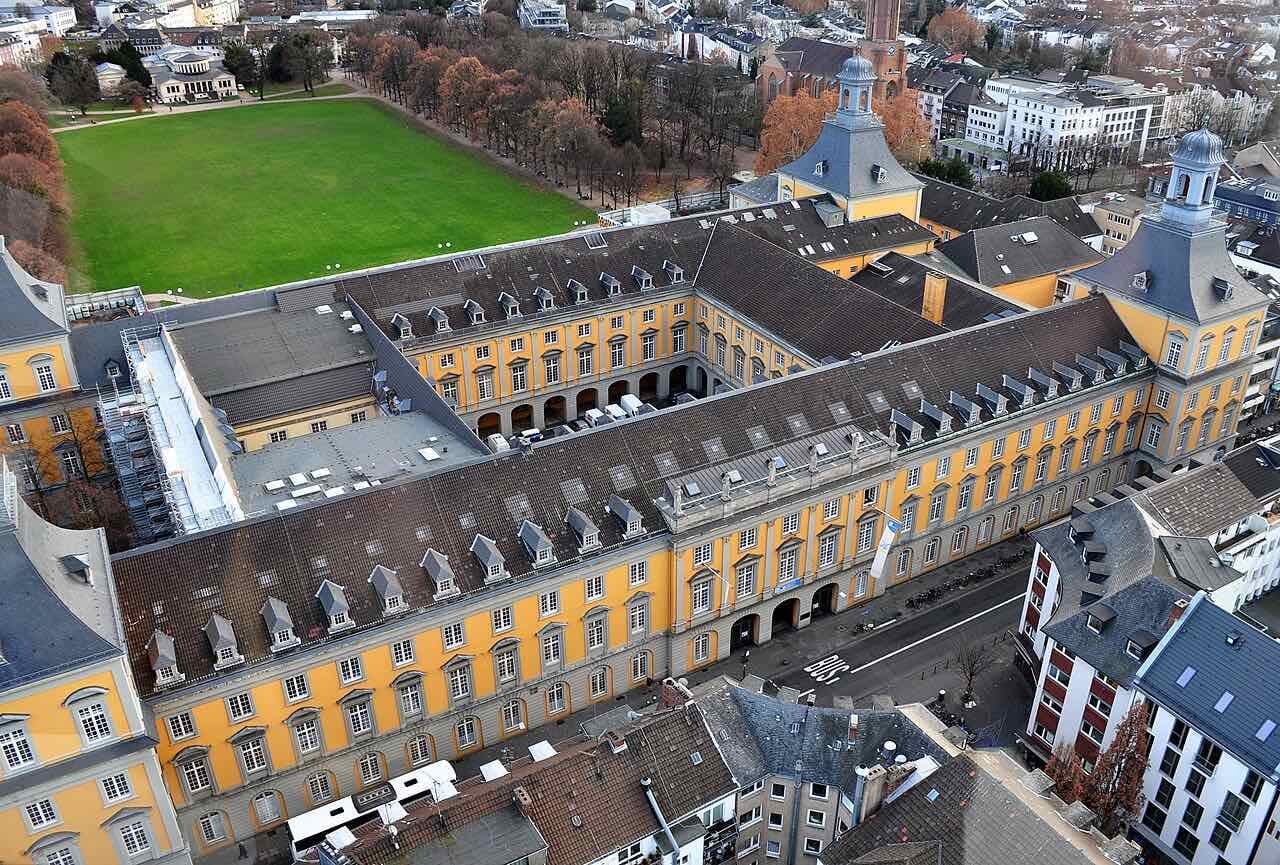 The long yellow main building of the University of Bonn seen from above, with the green lawn of the Hofgarten beside it